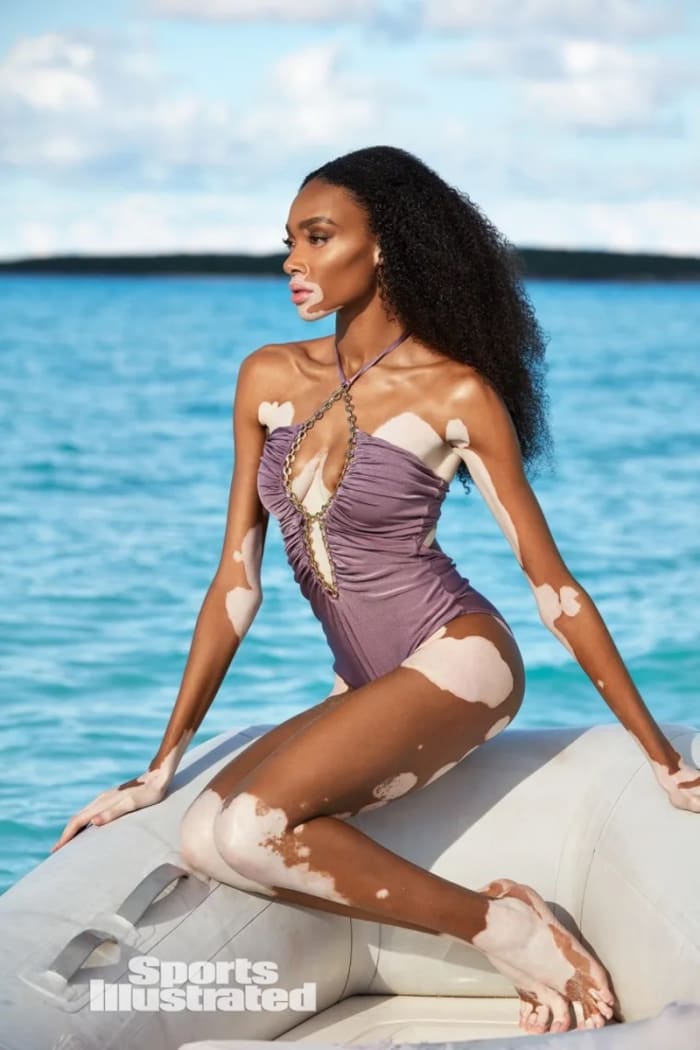 Winnie Harlow looks off into the distance, wearing a dusty lilac one-piece with gold ring detailing.